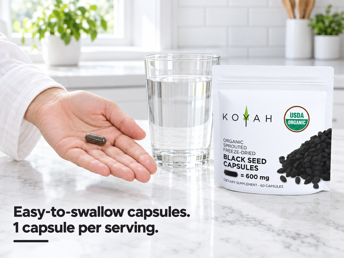 Hand holding a single KOYAH Organic Sprouted Black Seed Capsule next to the pouch and a glass of water: easy-to-swallow capsules, 1 capsule per serving